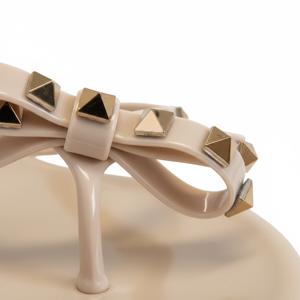 Close-up detail of gold pyramid studs and nude blush patent jelly bow on women's thong sandal