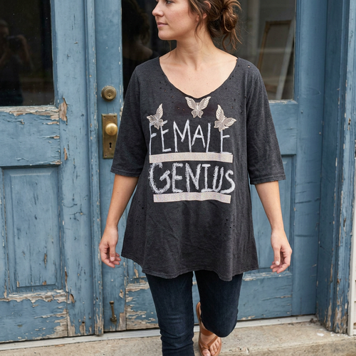 Woman in a charcoal Female Genius tee and dark jeans taking a step in front of a weathered blue door.