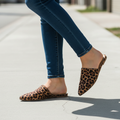 Woman walking on concrete sidewalk wearing Bella Marie leopard print pointed toe mule slides, mid-stride close-up from knees down