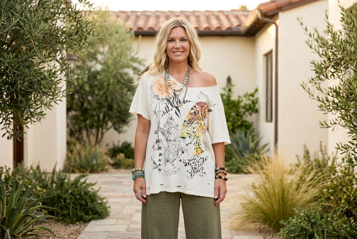 Woman standing in a sunny courtyard wearing an ivory oversized cat graphic tee off the shoulder with olive pants