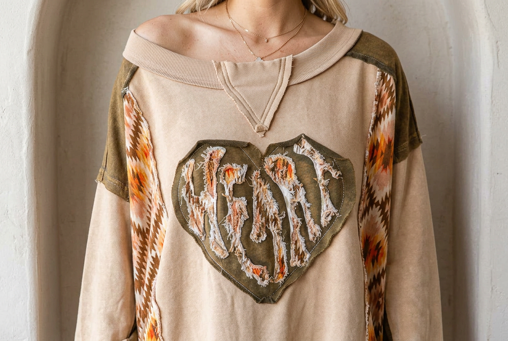 Front detail of a tan pullover with a large olive heart, frayed appliqué letters and patterned panels