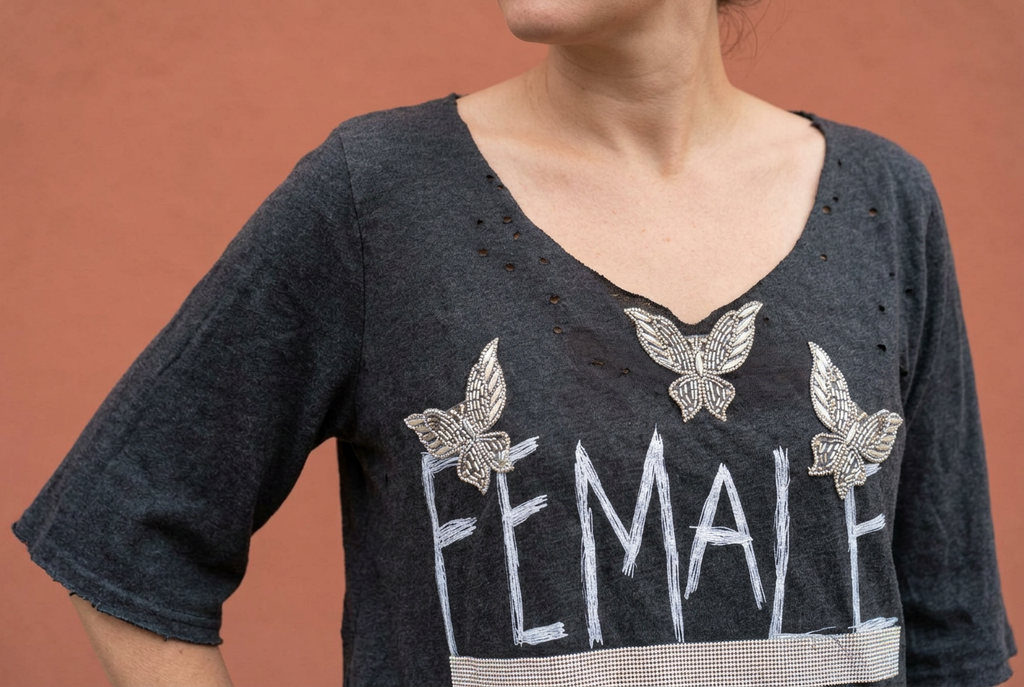 Angled detail of a charcoal tee’s distressed round neckline, wide sleeve, and beaded butterflies against a terracotta wall.