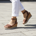 Woman walking in Cityclassified Junior-S brown cork wedge sandals on stone pavement wearing white linen trousers