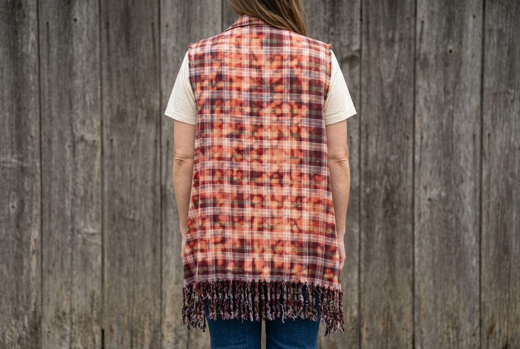 Rust plaid side of a long reversible vest, showing its back and fringed hem against weathered wood.