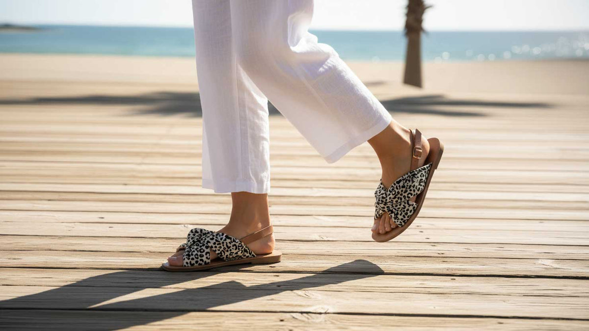 A person walking on a wooden boardwalk by the beach, wearing white pants and leopard-print slide sandals.
