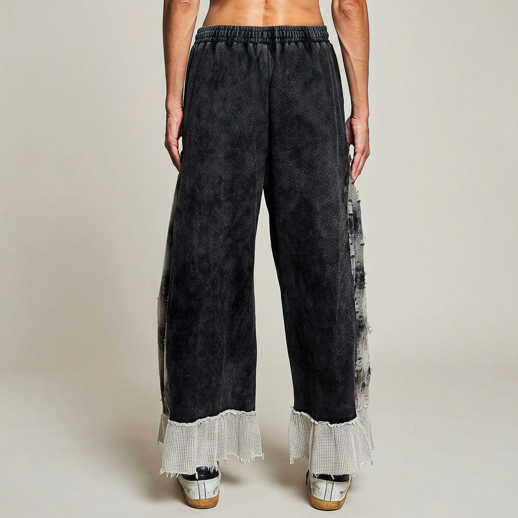 Back view of black wide-leg pants with plaid side inserts and pale textured ruffle hems on a warm-gray backdrop.