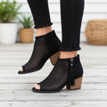 Black perforated peep-toe booties with fringe tassel detail, side profile on wooden deck steps in natural daylight