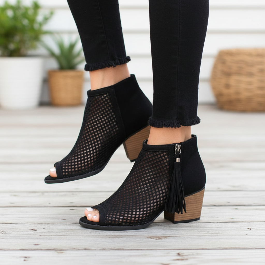 Black perforated peep-toe booties with fringe tassel detail, side profile on wooden deck steps in natural daylight