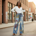 A woman stands on a city street wearing light blue wide-leg jeans with colorful embroidered and applique details, paired with a white long-sleeve blouse and brown heels.