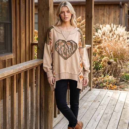 Relaxed tan heart pullover with an uneven hem, worn with black trousers on a wooden porch