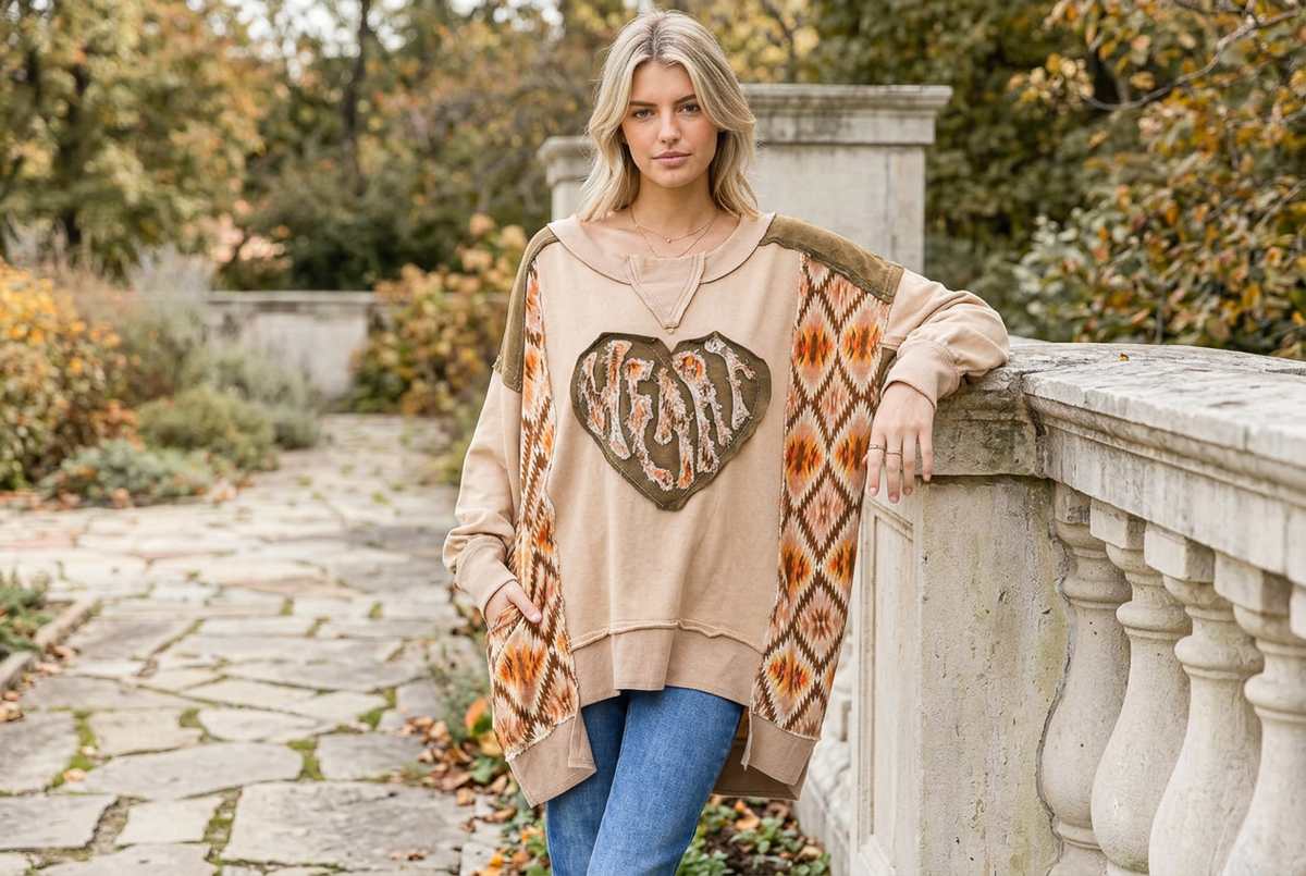 Tan heart patchwork pullover with dropped shoulders and a stepped hem, worn beside a stone garden wall