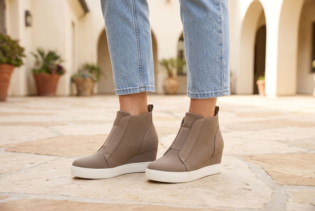 Woman in cropped jeans wearing taupe perforated wedge sneakers with white soles in a cream stucco courtyard.