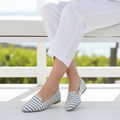 Grey and white stripe slip-on flats worn with cropped white linen trousers, seated outdoors in natural light