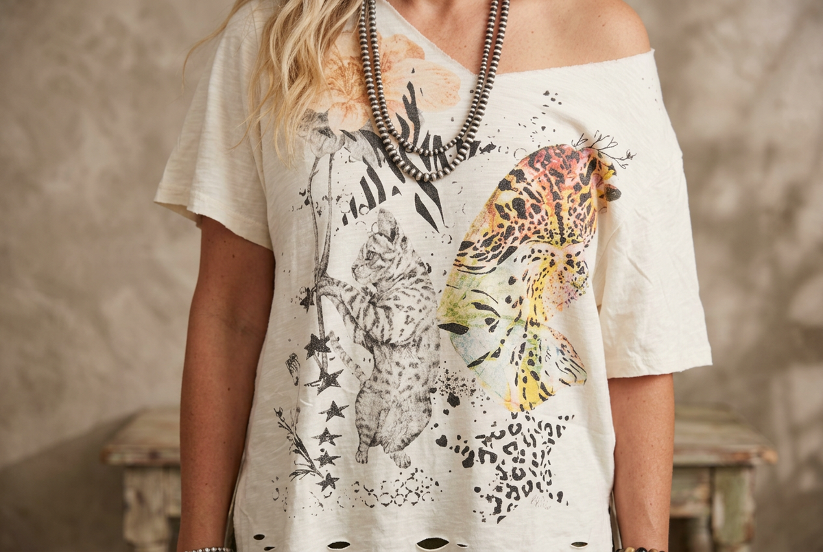 Front-detail crop of an ivory graphic tee showing the sketched cat and leopard-print butterfly wing with stars and splatter
