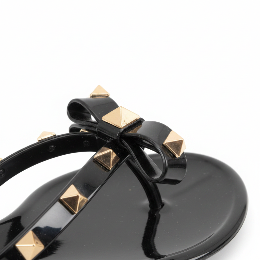 Close-up detail of gold pyramid studs and black patent jelly bow on women's thong sandal