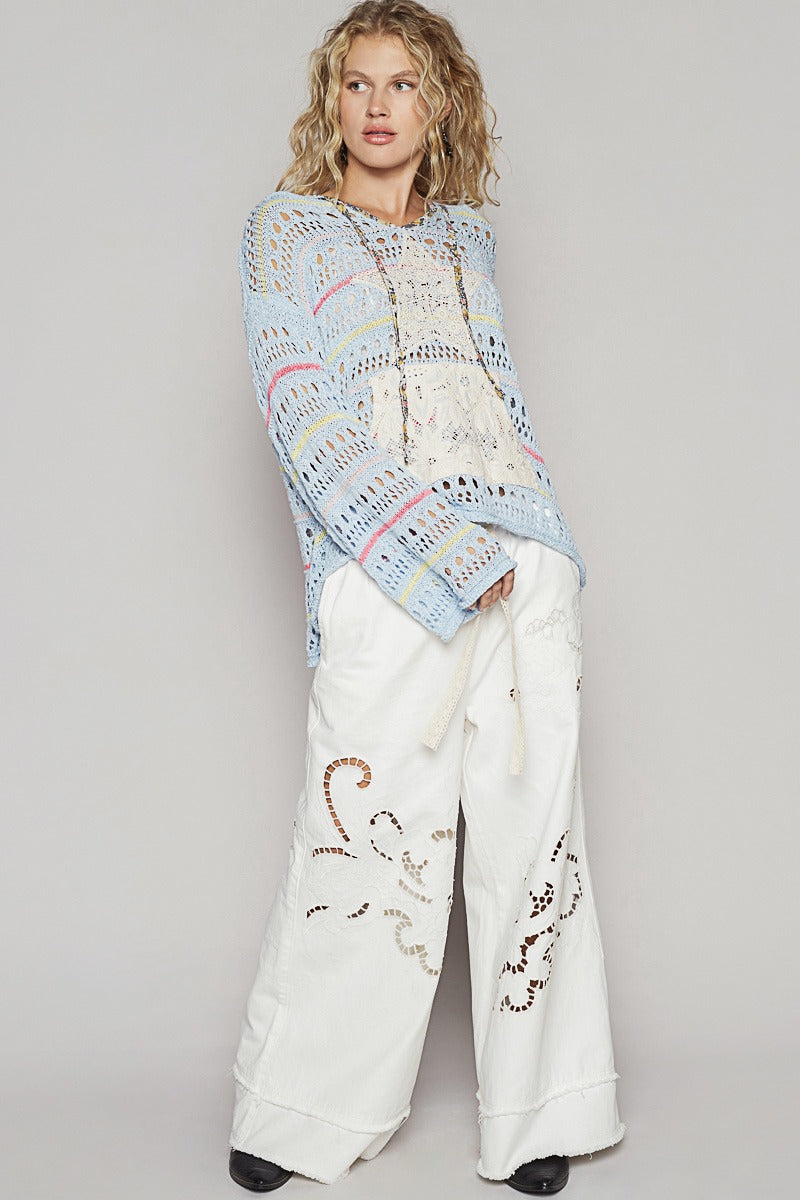 Woman wearing a colorful knitted poncho and white pants with decorative patterns on a plain background