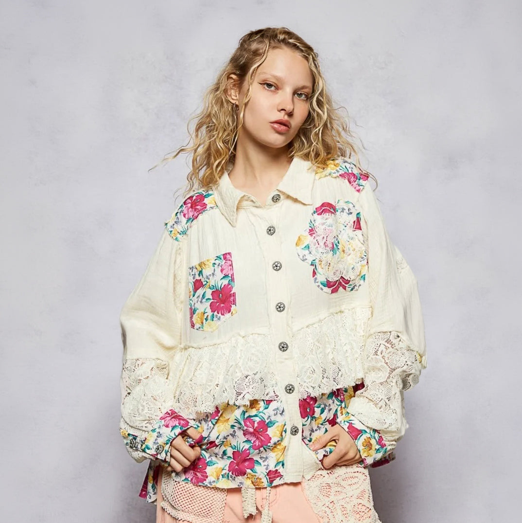 A model wearing a cream-colored patchwork blouse with colorful floral prints, lace ruffles, and button details, paired with light pink shorts, set against a textured gray studio background.