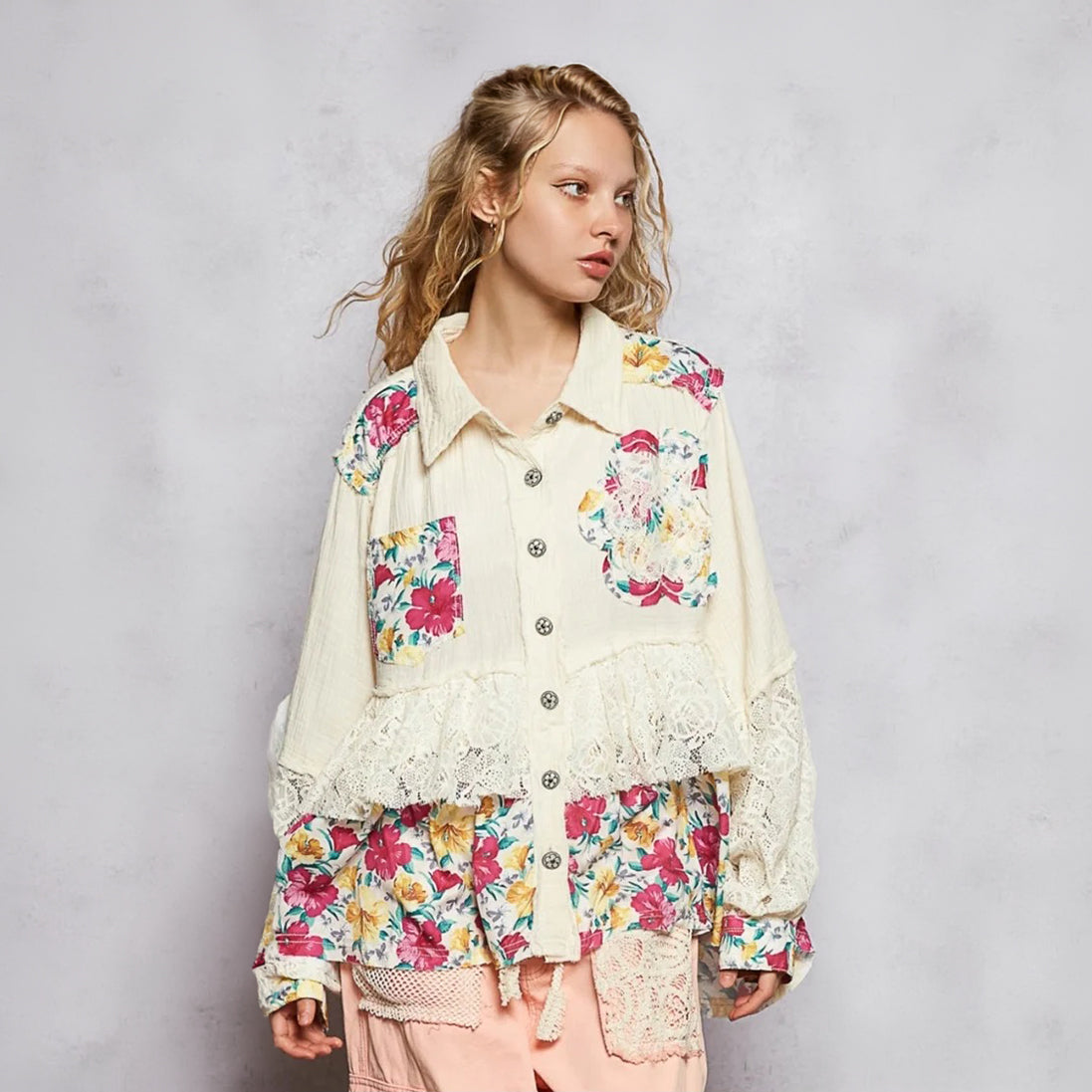 A woman wearing a cream-colored, layered button-up shirt with colorful floral patches, lace ruffles, and a drawstring hem, paired with light pink pants, set against a textured gray studio background.