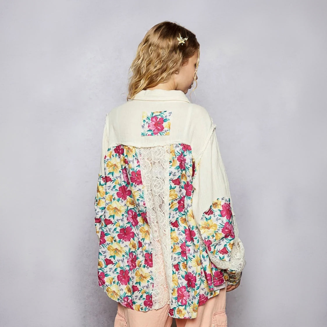 A woman shown from the back wearing a cream-colored patchwork jacket with vibrant pink, yellow, and blue floral patterns and a lace panel down the back, set against a plain gray studio background.
