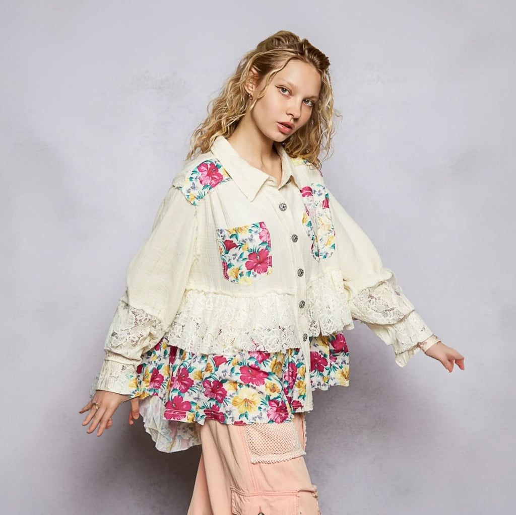 A model wearing a cream-colored, layered button-up shirt jacket decorated with colorful floral prints and lace details, paired with light pink cargo pants, set against a textured light gray studio background.