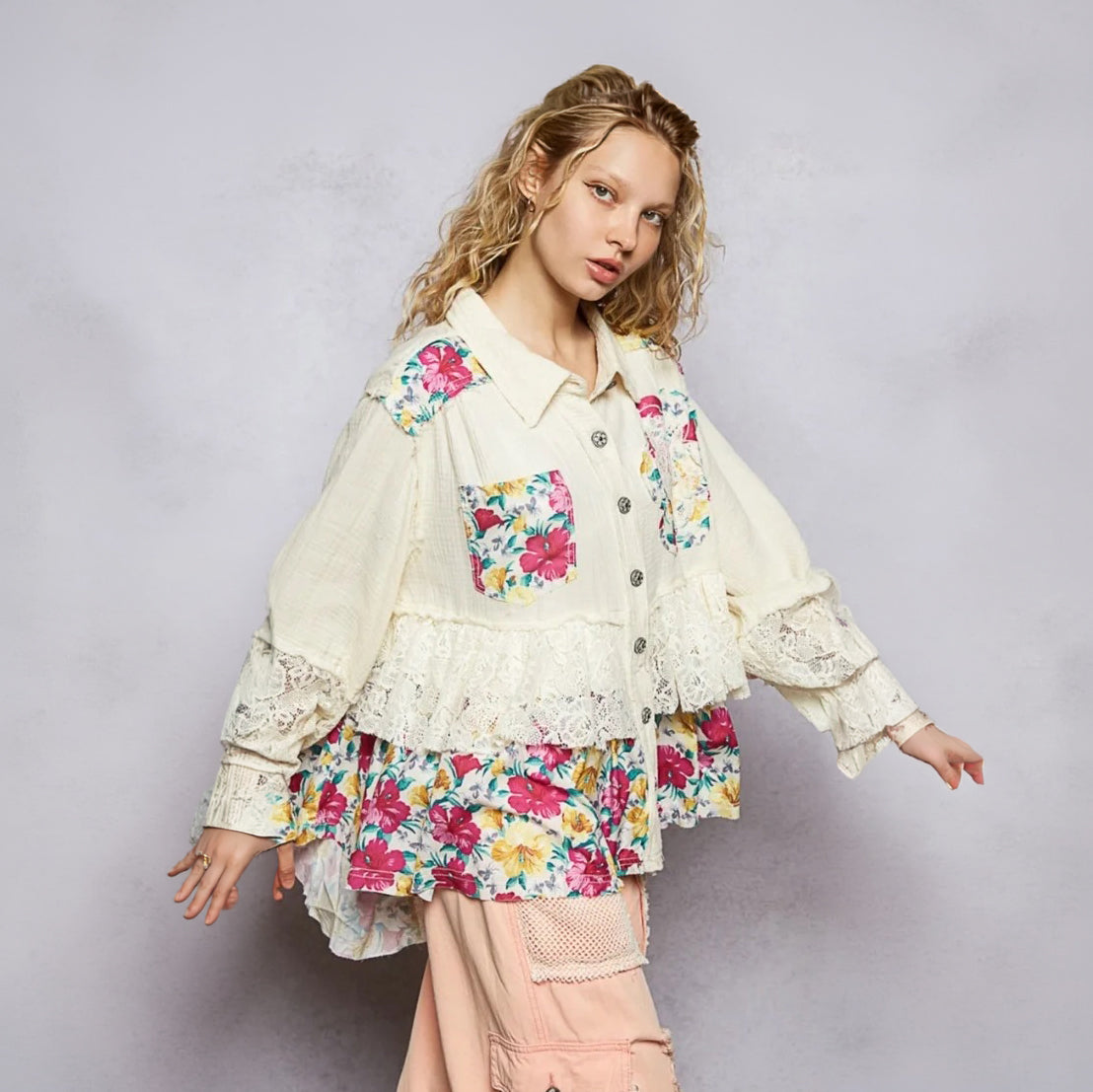A model wearing a cream-colored, layered button-up shirt jacket decorated with colorful floral prints and lace details, paired with light pink cargo pants, set against a textured light gray studio background.